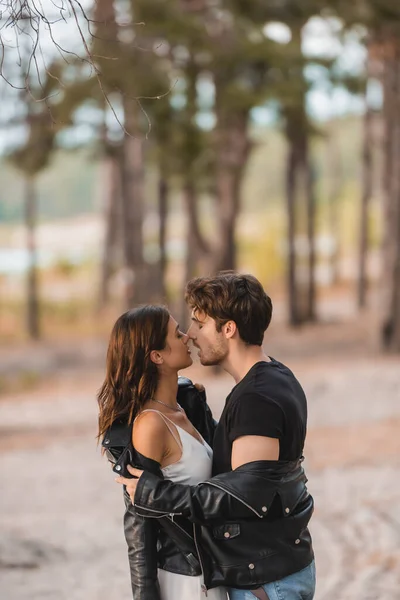 Vista lateral do homem beijando e tirando jaqueta de couro da namorada na floresta — Fotografia de Stock