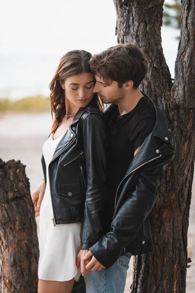 Selective focus of young couple in leather jackets holding hands near trees in forest — Stock Photo