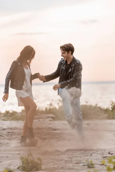 Selective focus of young couple in leather jackets throwing sand on beach — Stock Photo