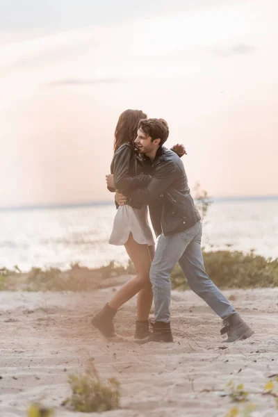 Selektiver Fokus des Mannes in Lederjacke umarmt Freundin im Kleid am Strand bei Sonnenuntergang — Stockfoto