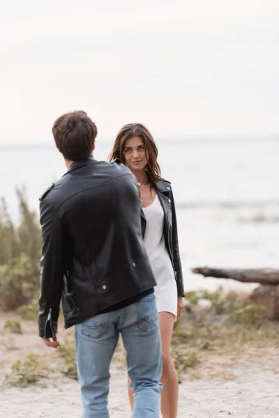 Selective focus of young woman in dress and leather jacket looking at boyfriend on beach — Stock Photo