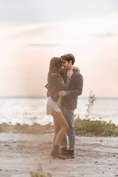 Selective focus of young man embracing girlfriend in dress and leather jacket on beach at sunset — Stock Photo