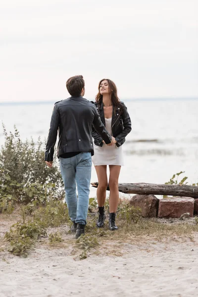 Brunette woman in dress and leather jacket holding hand of boyfriend on seaside — Stock Photo