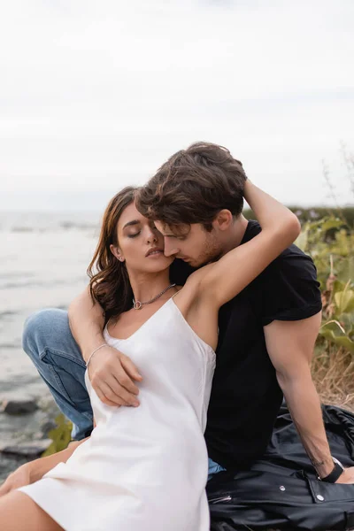 Mujer en vestido abrazando novio en chaqueta de cuero en la playa - foto de stock