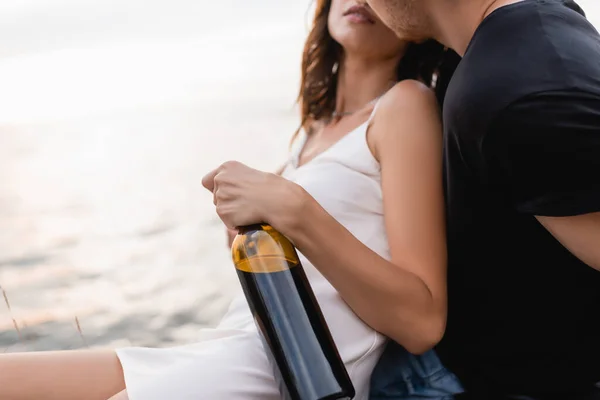 Cropped view of man kissing girlfriend in dress with bottle of wine near sea — Stock Photo