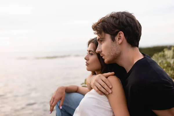 Young man embracing brunette girlfriend and looking at sea on beach — Stock Photo