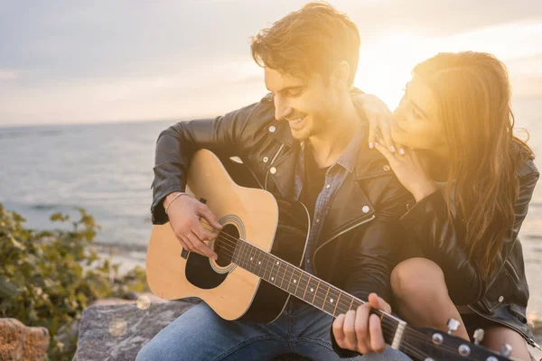 Jovem de jaqueta de couro tocando guitarra acústica perto da namorada na praia ao pôr do sol — Fotografia de Stock
