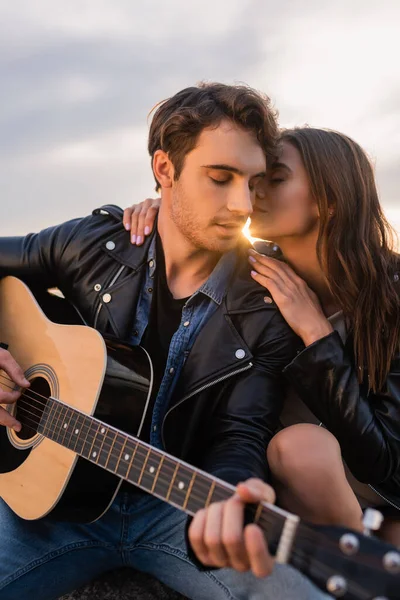 Selective focus of woman kissing boyfriend with acoustic guitar at sunset — Stock Photo