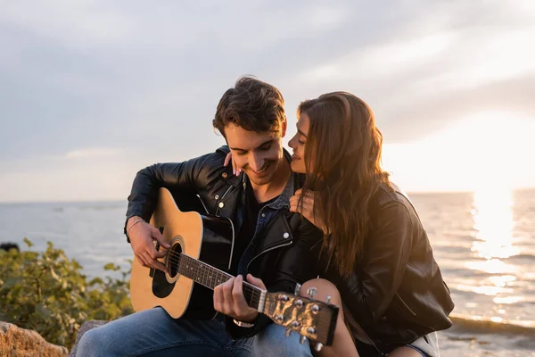 Foco seletivo da mulher em jaqueta de couro abraçando namorado tocando guitarra acústica durante o pôr do sol na praia — Fotografia de Stock