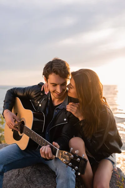 Foco seletivo da mulher abraçando namorado tocando guitarra acústica perto do mar durante o pôr do sol — Fotografia de Stock