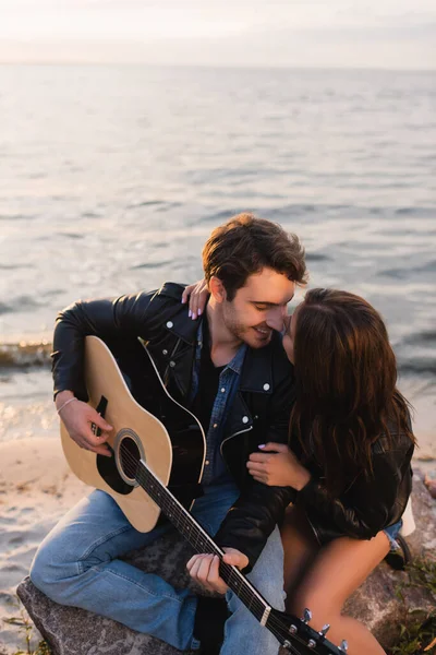 Pareja joven en chaquetas de cuero con guitarra acústica sentada en la playa durante el atardecer - foto de stock