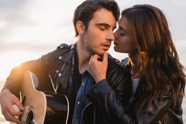 Жінка цілує хлопця в шкіряній куртці з акустичною гітарою під час заходу сонця. — стокове фото