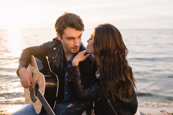 Mujer joven mirando novio con guitarra acústica en la playa al atardecer - foto de stock