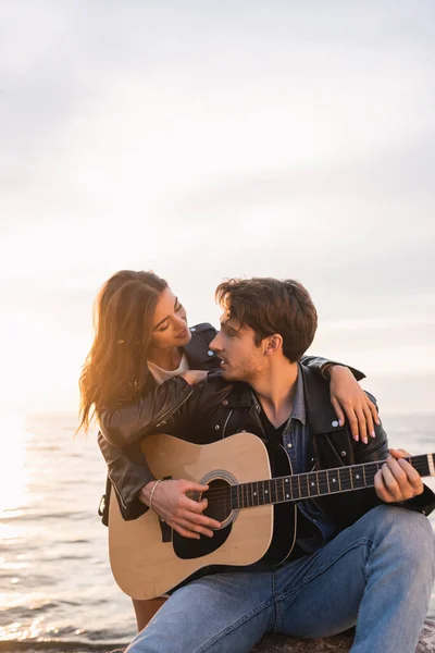 Jeune femme en veste en cuir embrassant petit ami jouant de la guitare acoustique près de la mer au coucher du soleil — Photo de stock