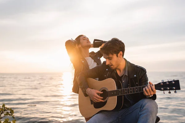 Foco seletivo do homem de jaqueta de couro tocando guitarra acústica perto da namorada durante o pôr do sol perto do mar — Fotografia de Stock
