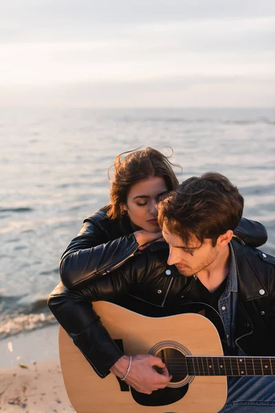 Mujer joven abrazando novio con guitarra acústica en la playa al atardecer - foto de stock