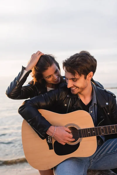 Burnette mulher tocando o cabelo e olhando para o namorado tocando guitarra acústica na praia à noite — Fotografia de Stock