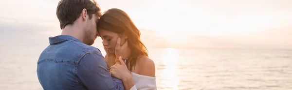 Tiro panorâmico de jovem tocando namorada com mar e pôr do sol céu no fundo — Fotografia de Stock