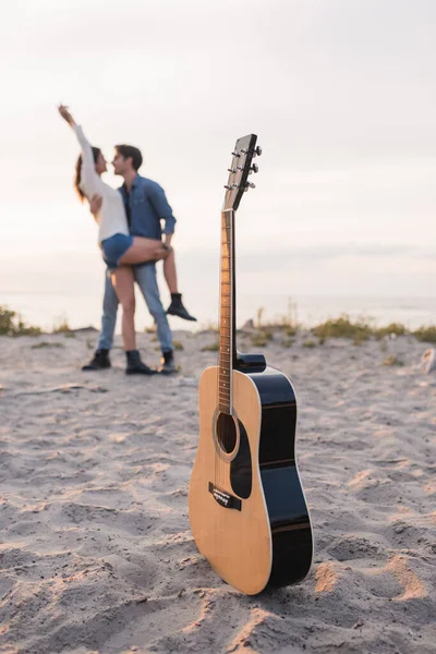 Selective focus of acoustic guitar on sand near young couple embracing on beach at sunset — Stock Photo