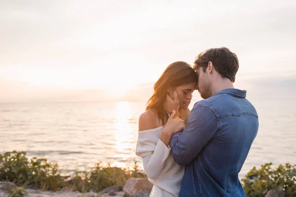 Joven tocando morena novia en la playa cerca del mar al atardecer - foto de stock