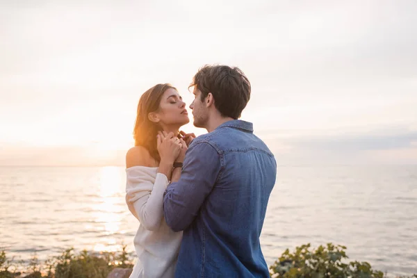Young man touching neck of brunette girlfriend on beach during sunset — Stock Photo