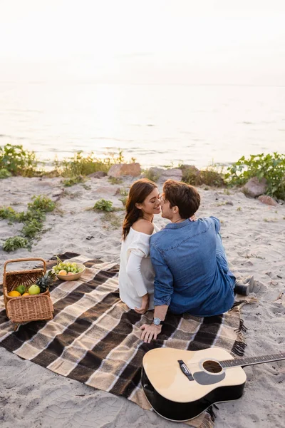 Pareja joven besándose cerca de la guitarra acústica y cesta de mimbre durante el picnic en la playa - foto de stock