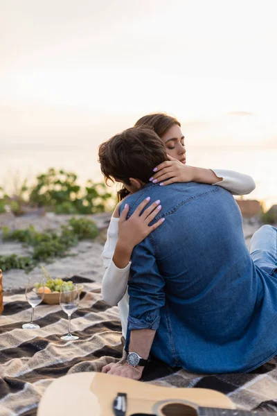 Selective focus of woman embracing boyfriend near acoustic guitar and wine glasses on beach — Stock Photo