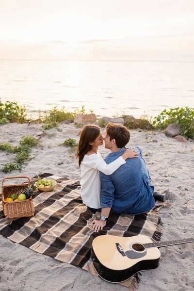 Selective focus of couple kissing near acoustic guitar during picnic on sea shore at sunset — Stock Photo