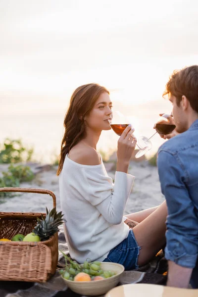Selektiver Fokus der Frau, die beim Picknick am Strand bei Sonnenuntergang Wein in der Nähe ihres Freundes trinkt — Stockfoto