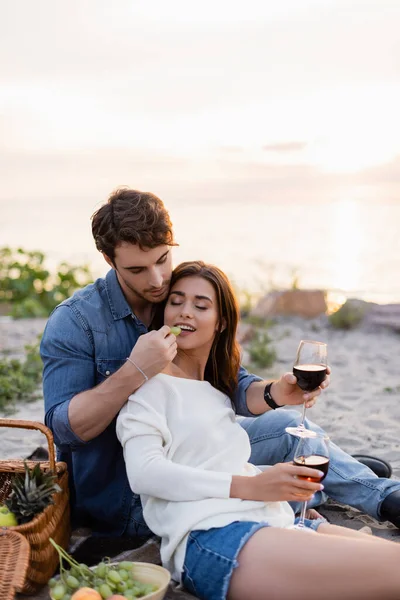 Focus selettivo dell'uomo che alimenta la ragazza con l'uva mentre tiene un bicchiere di vino sulla spiaggia — Foto stock