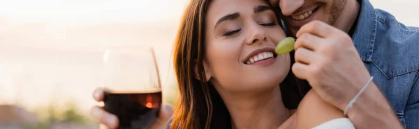 Website header of man with glass of wine feeding girlfriend with grape on beach at evening — Stock Photo