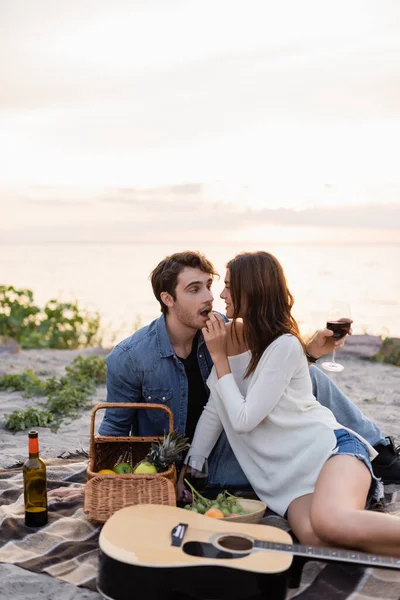 Selective focus of woman feeding boyfriend with grape near wine, fruits and acoustic guitar on blanket on beach — Stock Photo