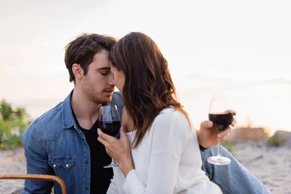 Молодая пара с бокалами вина целуется на пляже на закате — стоковое фото