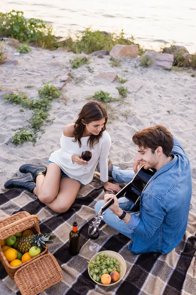 Vista ad alto angolo dell'uomo che suona la chitarra acustica vicino alla ragazza con un bicchiere di vino sulla spiaggia — Foto stock