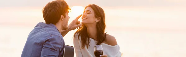 Colpo panoramico di uomo con chitarra acustica toccante naso di fidanzata con bicchiere di vino vicino al mare — Foto stock