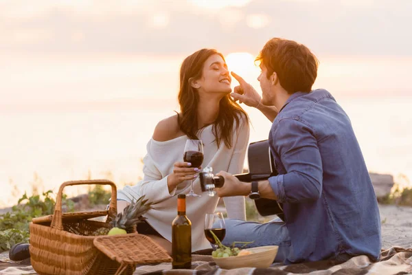 Чоловік з акустичною гітарою торкається носа дівчини зі склянкою вина на пляжі на заході сонця — стокове фото