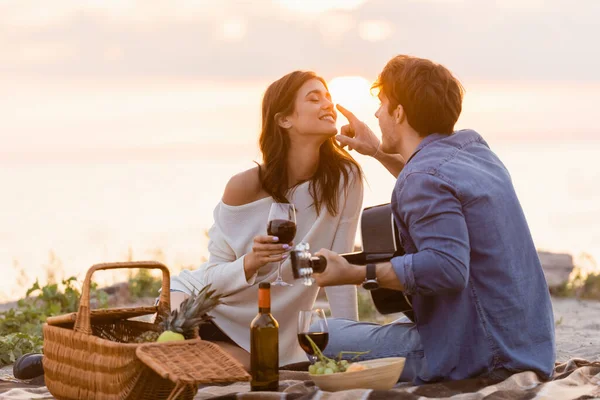 Hombre con guitarra acústica tocando nariz de novia con copa de vino en la playa al atardecer - foto de stock