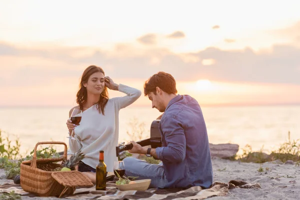 Paar spielt Akustikgitarre beim Picknick mit Wein am Strand bei Sonnenuntergang — Stockfoto