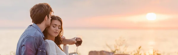 Tiro panorâmico de mulher com copo de vinho abraçando namorada na praia ao pôr do sol — Fotografia de Stock