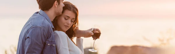 Encabezado del sitio web de hombre abrazando a la mujer mientras sostiene una copa de vino en la playa al atardecer - foto de stock
