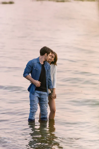 Jovem casal de mãos dadas em pé na água do mar ao pôr do sol — Fotografia de Stock