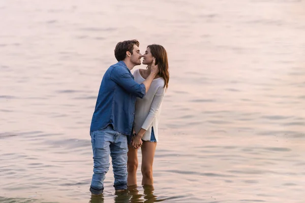 Giovane uomo baciare fidanzata mentre in piedi in mare al tramonto — Foto stock