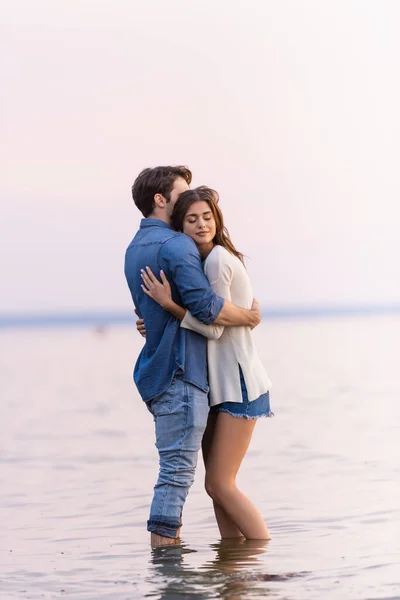 Side view of brunette woman with closed eyes hugging boyfriend while standing in sea — Stock Photo