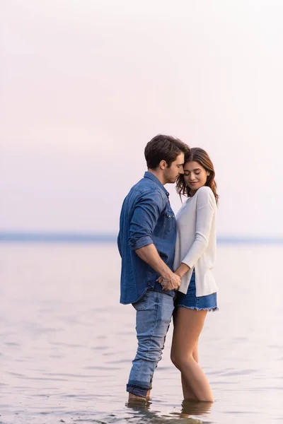 Side view of young couple holding hands while standing in sea at evening — Stock Photo