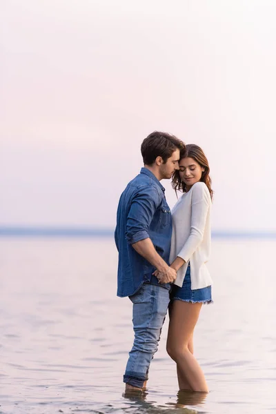 Vue latérale du jeune couple tenant la main debout en mer le soir — Photo de stock