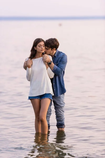 Man hugging brunette girlfriend with closed eyes while standing in sea at sunset — Stock Photo