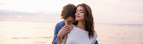 Vue panoramique de l'homme touchant petite amie brune avec les yeux fermés près de la mer au coucher du soleil — Photo de stock