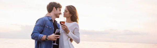 Panoramaausrichtung der Frau, die ihren Freund umarmt, während sie ein Glas Wein in der Nähe des Meeres hält — Stockfoto