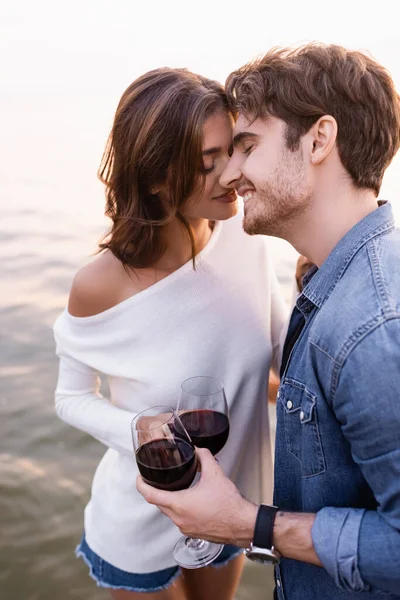 Joven con copa de vino de pie cerca de la novia y el mar al atardecer - foto de stock