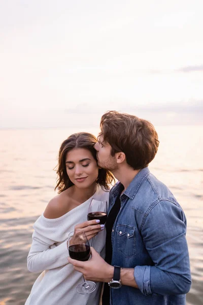 Young man kissing girlfriend with glass of wine near sea at evening — Stock Photo